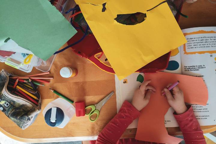 A birdseye view of a child&#039;s hands drawing on paper. They are surrounded by arts and crafts supplies such as coloured pencils, scissors and a gluestick.