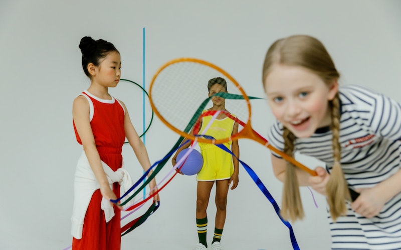 Three young children in white sports hall holding tennis rackets and playing together.