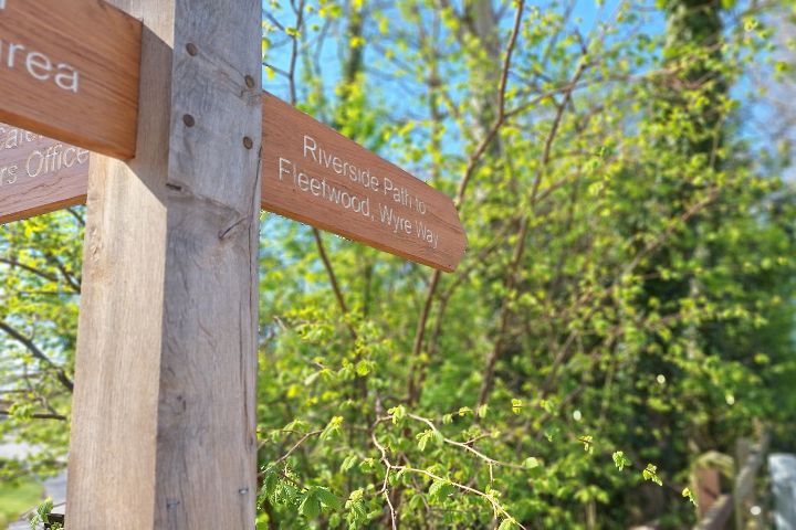 A wooden sign post pointing to Riverside Path to Fleetwood, Wyre Way amongst the trees.