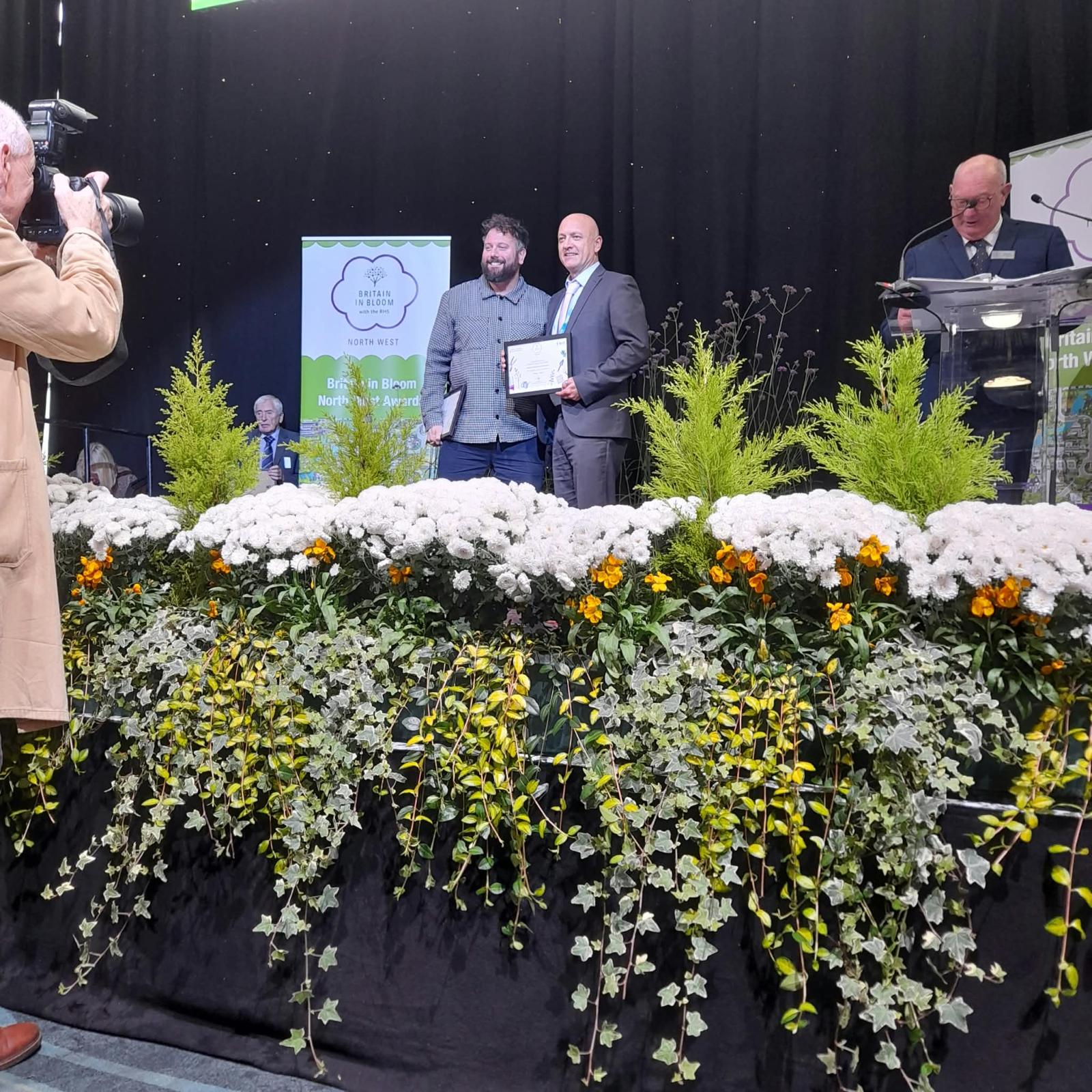 Two men are on stage accepting an award certificate, smiling and posing for a photographer.