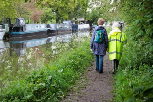 Two people walking along a canal with canal boats on the far side