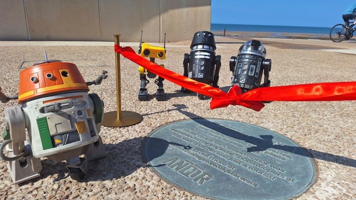A minature sized scene of three robots from the movie Star Wars standing next to a red ribbon held between two metal pillars above the new Andor plaque on Cleveleys beach.