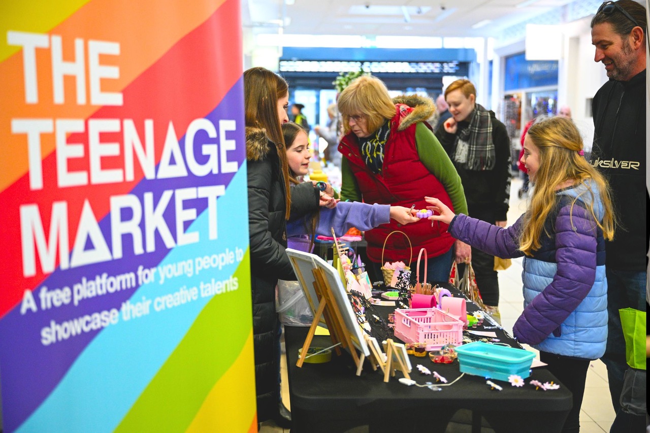 A colourful sign that says 'The Teenage Market' with young people browsing stalls.
