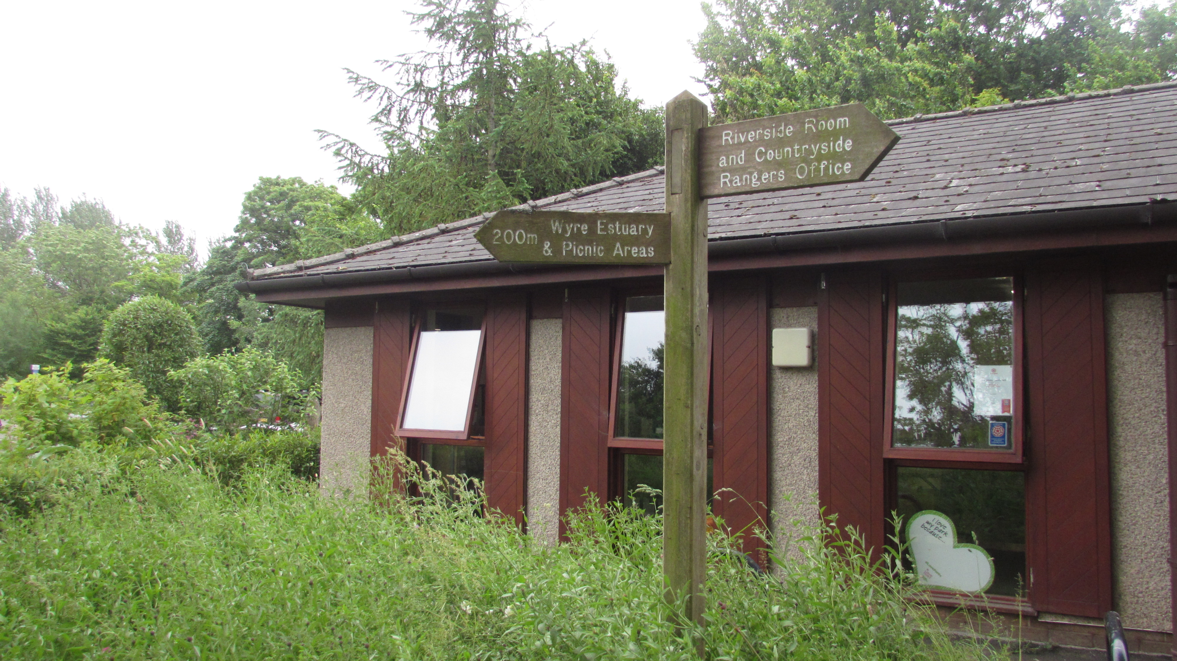 A building at a country park. A signpost in front of the building directs to the Riverside Room and Rangers Office in one direction, and the Wyre Estuary in the opposite direction.