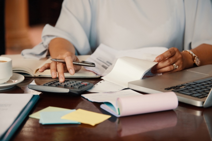 Person using a calculator. On the table their is a laptop, coffee, and scattered paper.