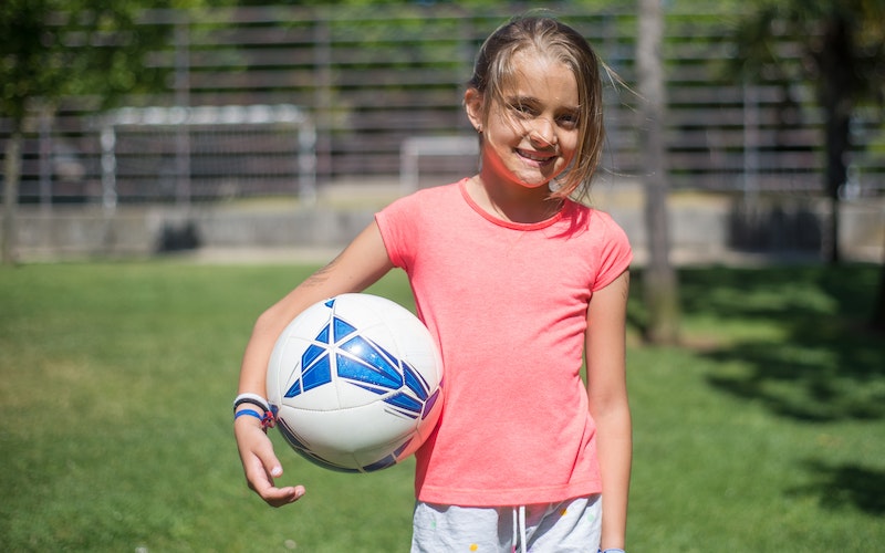 Young girl smiling for camera, holding a football whilst stood on a football field.