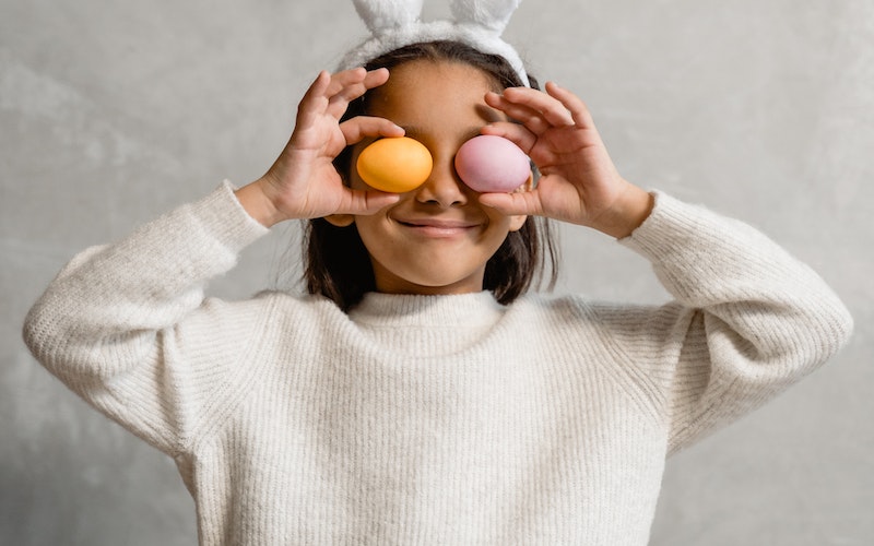 Young girl wearing rabbit ears and holding painted boiler eggs over her eyes for Easter.